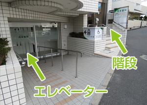 受付は2Fです。右手の階段か、スロープを下りてエレベーターでお上がりください